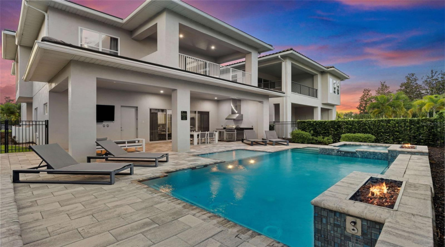 This resort-style backyard is a private oasis with a heated custom pool, raised spa, paver deck, lush landscaping, and striking fire bowls. A covered lanai with a summer kitchen and outdoor TV, plus a second-story balcony overlooking the pool, create the perfect setting for relaxation and entertaining from day to night.