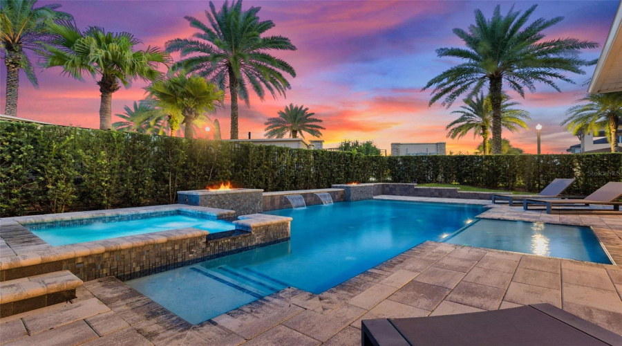 This resort-style backyard is a private oasis with a heated custom pool, raised spa, paver deck, lush landscaping, and striking fire bowls. A covered lanai with a summer kitchen and outdoor TV, plus a second-story balcony overlooking the pool, create the perfect setting for relaxation and entertaining from day to night.