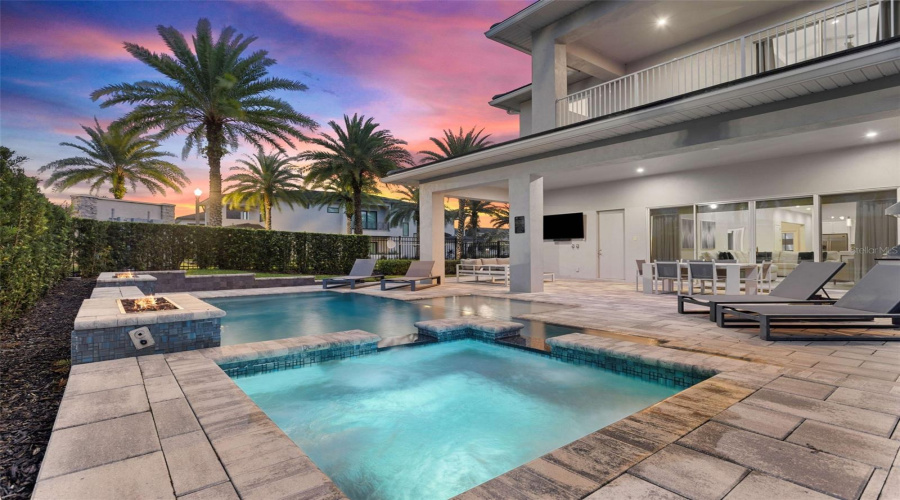 This resort-style backyard is a private oasis with a heated custom pool, raised spa, paver deck, lush landscaping, and striking fire bowls. A covered lanai with a summer kitchen and outdoor TV, plus a second-story balcony overlooking the pool, create the perfect setting for relaxation and entertaining from day to night.