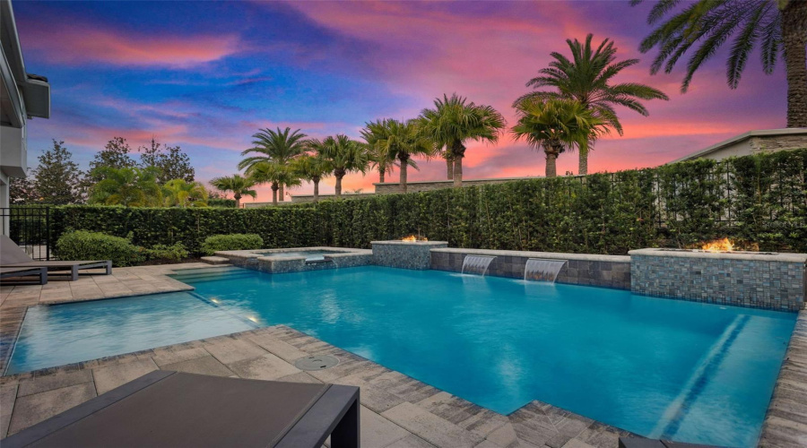 This resort-style backyard is a private oasis with a heated custom pool, raised spa, paver deck, lush landscaping, and striking fire bowls. A covered lanai with a summer kitchen and outdoor TV, plus a second-story balcony overlooking the pool, create the perfect setting for relaxation and entertaining from day to night.