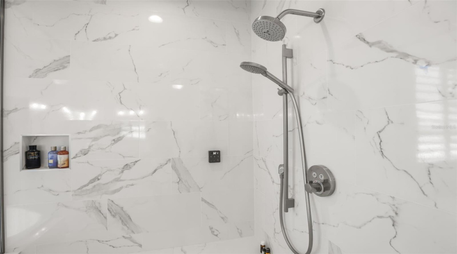 The oversized shower showcases marble-look tile, built-in niches, a bench, and multiple shower fixtures for an upgraded bathing experience.