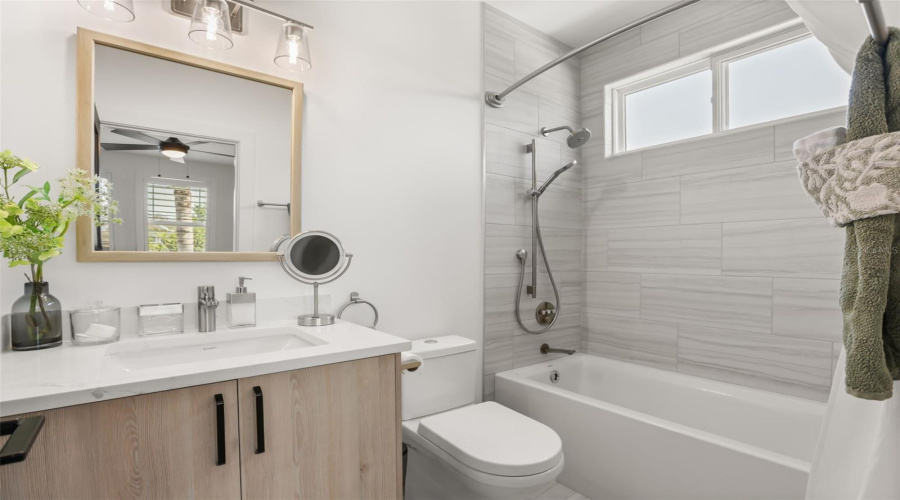 The bathroom includes a tub-shower combination with marble-look tile, a light wood vanity, and a window that brings in natural light.