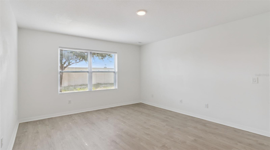 Sunlit Bedroom:Well-lit bedroom showcasing light wood-grain vinyl flooring, fresh neutral paint, and a large window with fitted horizontal blinds letting in abundant natural light.