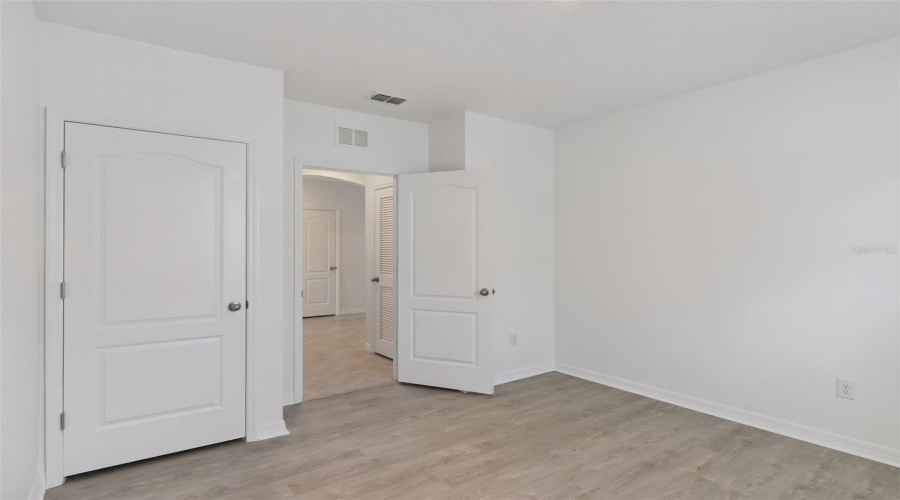 Bedroom & Closet Entry:Clean bedroom perspective highlighting wood-look flooring, white trim, built-in closet door, and open doorway leading to the main tiled hallway.