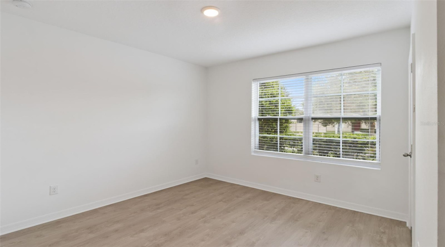 Bedroom Window Corner:Inviting bedroom corner featuring warm wood-look flooring, pristine white walls, and a large picture window with blinds overlooking exterior landscaping.