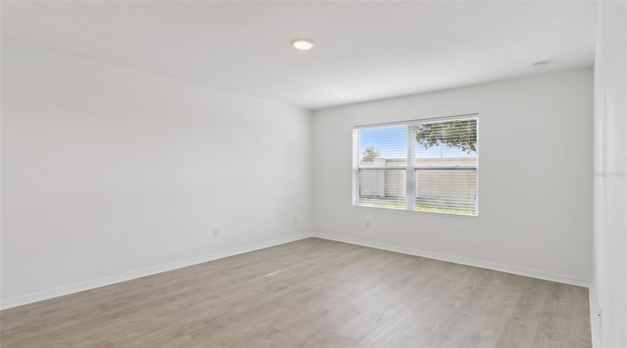 Primary Bedroom Suite:Spacious primary suite boasting light wood-grain vinyl flooring, recessed ceiling lighting, clean white walls, and a wide window letting in abundant natural light.