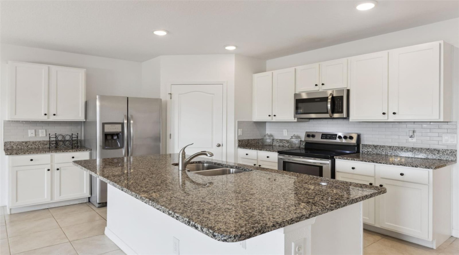 Full Kitchen View:Spacious kitchen features a generous prep island, mottled granite countertops, white cabinetry, subway tile backsplash, stainless steel appliances, and low-maintenance tile flooring.