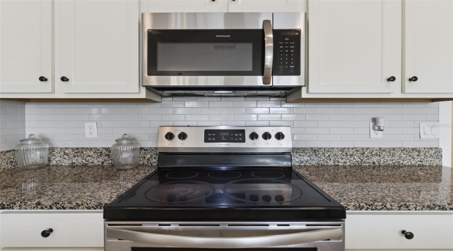 Cooking Station:Modern cook station equipped with a stainless steel smooth-top electric range, built-in microwave, crisp white subway tile backsplash, and granite counters.