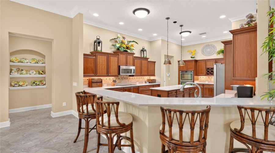 Overview Of The Gorgeous Kitchen - Shows Staggered Height Wood Cabinets (Cherry Stain) W/ Crown &Amp; Light Chair Rail Molding, Under Cabinet Lighting, Pull Outs, And A Built-In Pantry; Tile Backsplash; Quartz Counters; A Large Island W/ Pendant Lighting; Raised Peninsular Breakfast Bar; And Ss Appliances, Including A Built-In Double Oven, Cooktop, Microwave Hood, Bosch Dw (2025) &Amp; French-Door Refrigerator (2024). Note Hallway To L Of Kitchen With Niche Serves As Entryway To Primary Suite (L Turn) And To Interior Laundry (R Turn).