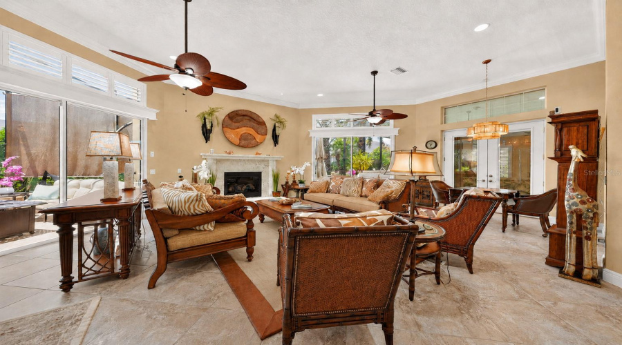 View Of The Center Section Of The Gathering Room - The Impressive Fireplace W/ A Cut Coral Surround &Amp; Mantle Is The Focal Point Of This Conversation/Seating Area. Upscale Furniture Pieces Manufactured By Tommy Bahama Home Included Are: 1) Two Rattan &Amp; Carved Wood Sofas W/ Tropical Decorative Pillows W/ Zebra Stripes/Large Leaves; 2) Two Accent Chairs Made Of Rattan/Bamboo Frame With Tropical Leaf-Pattern Cushions; 3) A Side Table W/ Round Wood Pedestal, Glass Top, And Rattan Base And 4) Rectangular Wood-Plank Top Coffee Table W/ Woven Rattan Apron Drawer/Storage &Amp; Turned Bun Feet, And 5) Nassau Console/Sofa Table, Part Of Island Estate Collection.