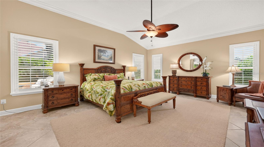Primary Suite - Wide Angle View Of The Bedroom From The French Doors Shows A Vaulted Ceiling W/ Crown Molding, Ceiling Fan And Multiple Windows With Plantation Shutters. Note Tommy Bahama Palmiers Green King Comforter Set On Tempurpedic Adjustable Bed.