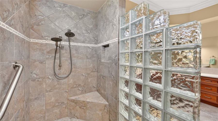 Primary Ensuite Bath - Interior View Of Roman Shower Highlighting The Tiled Walls With Decorative Tile Accent Band, Built-In Corner Seat, Recessed Shower Niche, Grab Bar, And Multiple Shower Heads, Including A Handheld Type.