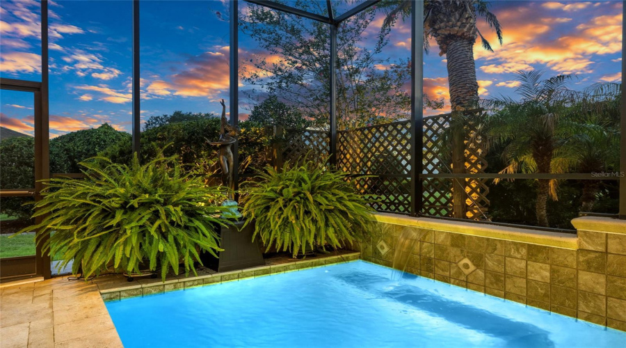 South End Of Pool Area @ Twilight/Dusk - Close-Up View Of A Cascading Waterfall Flowing From The Raised Decorative Tiled Wall Into The Pool. Potted Ferns, Tropical Landscaping, And Lattice Privacy Panels Along The Perimeter Of Mansard Screen Enclosure Produces A Feeling Of Serene Privacy.
