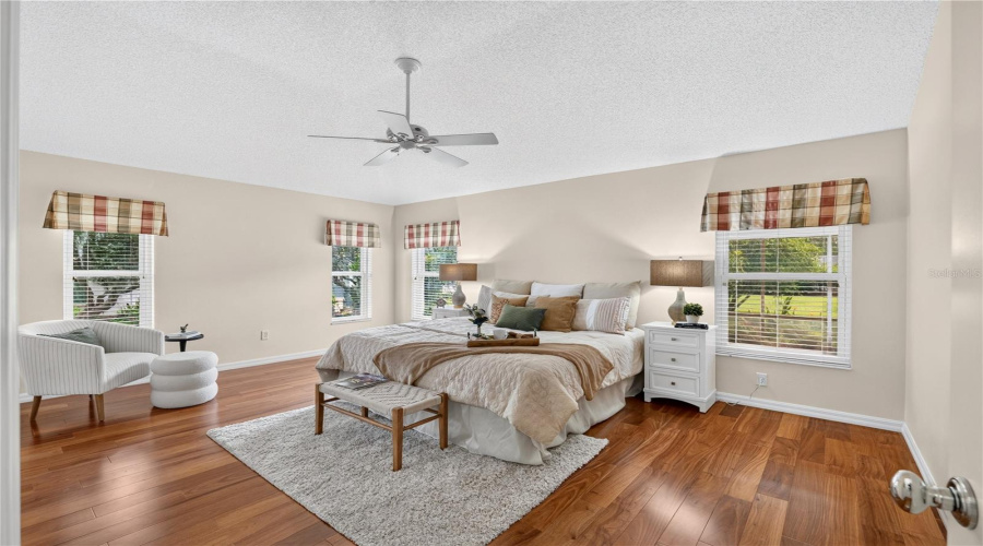 Spacious Primary Bedroom With High Ceilings, Rich Wood Flooring, Ceiling Fan, And Multiple Windows For Natural Light.