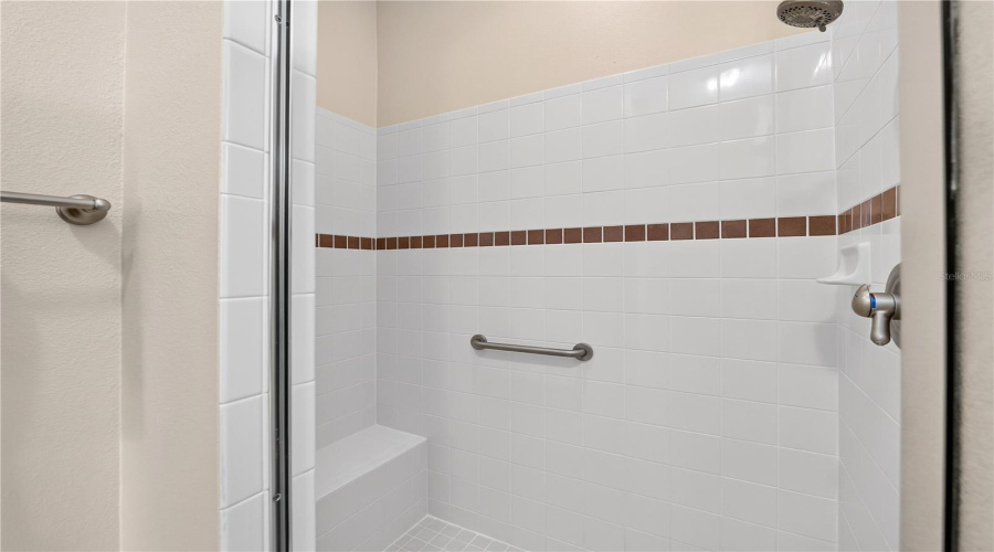 Primary Shower With Neutral Tile Surround, Built-In Bench, Grab Bar, And Decorative Accent Detail.