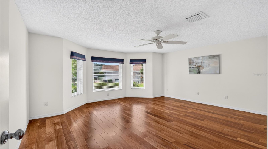 Spacious Guest Bedroom With Wood Flooring, Ceiling Fan, And A Bay-Style Window Area Bringing In Natural Light.
