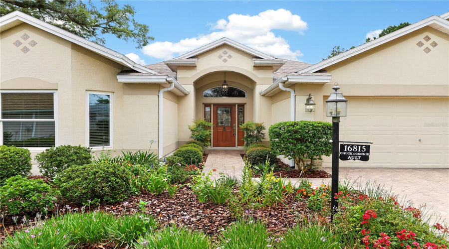 Inviting Front Entry Featuring An Arched Covered Entrance, Glass Front Door, And Lush Garden Beds.