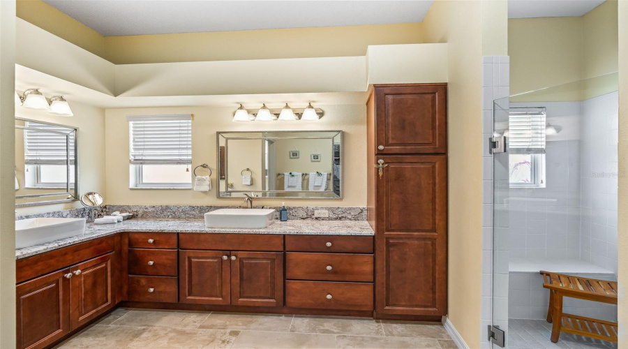 The Primary Bathroom Offers Extensive Wood Cabinetry, A Tall Linen Cabinet, Dual Vanity Areas, And Multiple Windows For Natural Light.