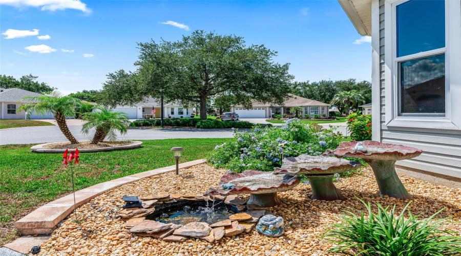 Charming Landscaped Garden Area With Mature Plantings, Decorative Stone Accents, And A Peaceful Water Feature Creates An Inviting Outdoor Focal Point.