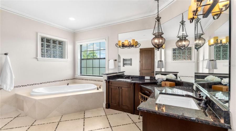 This Second Primary En-Suite Bath Pairs A Jacuzzi Soaking Tub With Custom Cabinetry, Natural Stone Surfaces And Elegant Chandelier Lighting For A Refined Private Retreat.