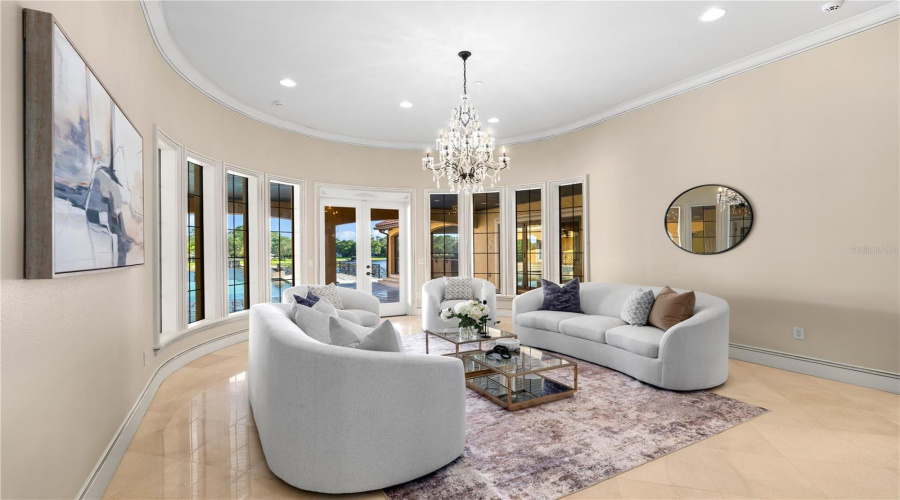 The Formal Living Room Is Enveloped By A Graceful Curved Wall Of Windows, Creating A Luminous, Sophisticated Setting Beneath An Elegant Crystal Chandelier.