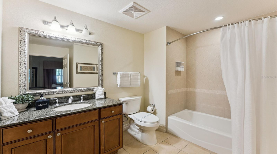 Second Full Bathroom: Guest Bathroom Featuring A Wood Vanity With Granite Countertop, Decorative Framed Mirror, Toilet, And Tiled Shower/Tub Combination.