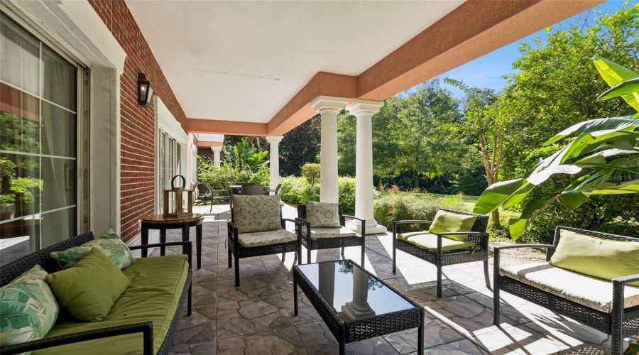 Covered Patio Seating: Covered Outdoor Patio With Stone-Patterned Tile Flooring, Classic Red Brick Wall Accents, Comfortable Wicker Lounge Seating, And Views Of Surrounding Tropical Flora.