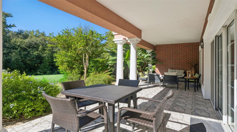 Outdoor Dining Area: Spacious Covered Patio Section Featuring A Four-Person Outdoor Wicker Dining Set, White Support Columns, Sliding Glass Door Access, And Lush Greenery Surroundings.