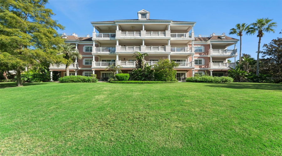 Rear Building Elevation: Full Rear Elevation View Of The Multi-Story Condominium Building Featuring Multi-Tiered Covered Balconies, A Sweeping Green Lawn, And Tropical Landscaping.