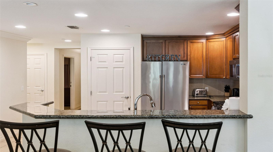 Breakfast Bar &Amp; Kitchen: Raised Granite Breakfast Bar Ideal For Casual Seating, Featuring A Stainless Steel Refrigerator, Recessed Lighting, And A Pantry Door.