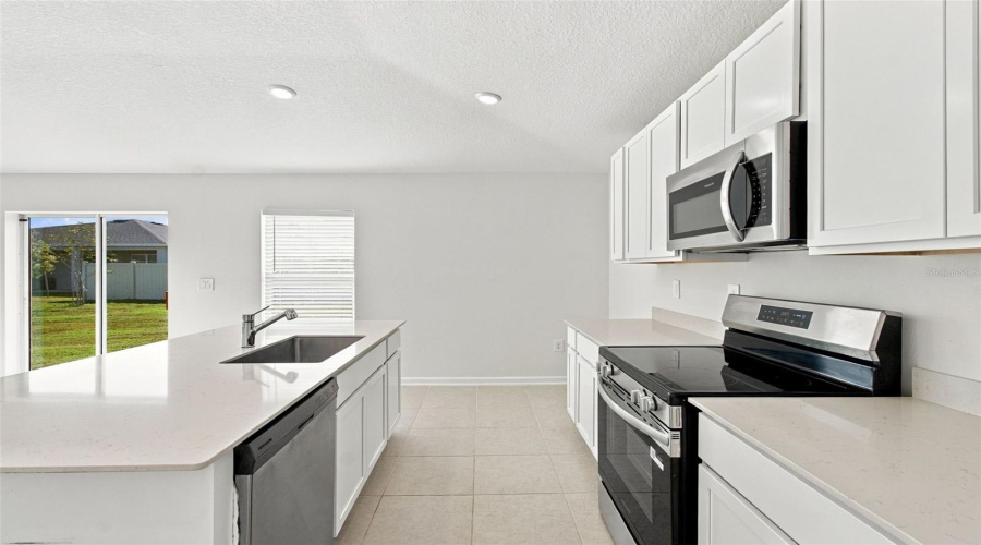 The Kitchen Offers A Spacious Island With Breakfast Bar, White Shaker-Style Cabinetry, And Quartz Countertops, All Overlooking The Dining And Living Areas.